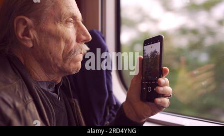 Elderly man traveling by train and taking pictures of the landscape through the window using a smartphone Stock Photo
