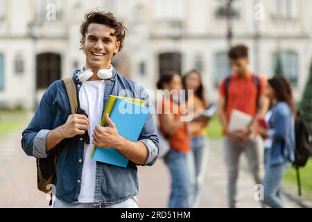 Handsome Young Male Student With Workbooks In Hands Posing Outdoors At Campus Stock Photo