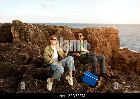 Happy Young Man And Woman Relaxing On Rocks Near Ocean Shore Stock Photo
