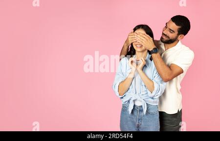 Loving handsome guy covering excited woman eyes, pink background Stock Photo