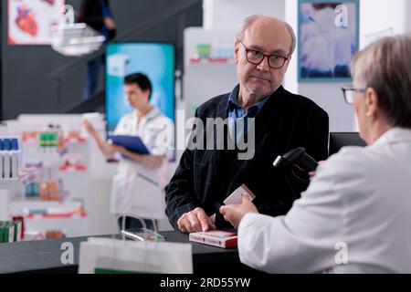 Elderly customer wating for senior cashier checking supplements price at pharmacy store checkout. Old man shopping in drugstore, buying healthcare vitamin and standing near counter desk Stock Photo