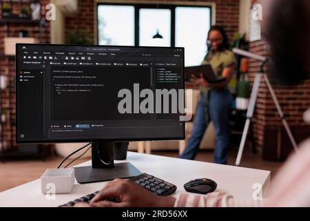 Freelance programmer developing source code on personal computer while girlfriend is using laptop standing in living room. African american couple working remote programming software together. Stock Photo
