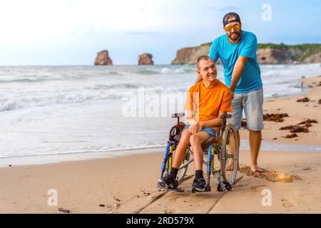 A disabled person in a wheelchair on the beach pushed by a friends by the sea Stock Photo