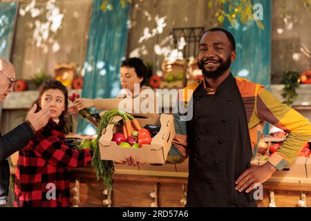 Cheerful small business owner posing at farmers market, wearing apron looking at camera. Male food marketplace vendor selling fresh organic produce and standing at local market. Stock Photo