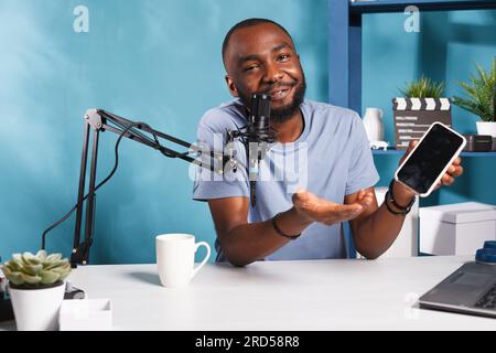 Blogger reviewing new smartphone, speaking in microphone and recording video for internet channel. African american smiling blogger holding mobile phone and streaming for social media Stock Photo