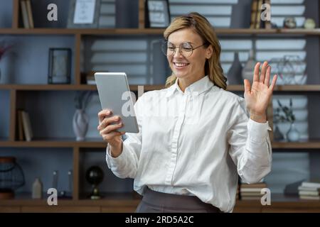 Mature experienced business woman boss using tablet computer for video call, senior woman working from home office remotely, smiling talking to colleagues at remote meeting. Stock Photo