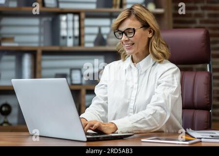 Mature smiling businesswoman working in evening inside home office, woman using laptop at work, typing on keyboard smiling, remote work from home. Stock Photo