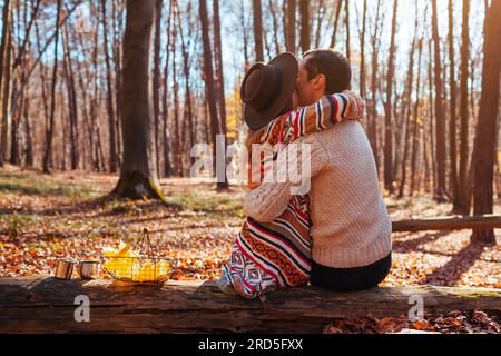 Back view of couple hugging in autumn forest. Man and woman sitting on tree trunk enjoying fall landscape on date. Young people kissing Stock Photo