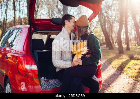 Happy loving couple enjoying date in car trunk hugging in autumn forest. Man and woman travel by auto. Young woman holding yellow leaves Stock Photo