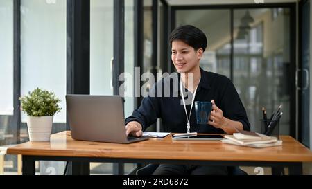 A happy and handsome millennial asian businessman or male office worker in casual clothes working on his work on his laptop at his desk in the office. Stock Photo