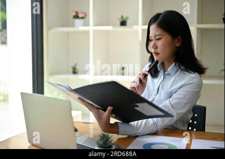 A stressed and focused Asian businesswoman or female manager reading and examining business documents at her desk in her office. Stock Photo