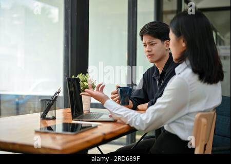 Two serious and professional Asian office workers are working remotely at a coffee shop together, discussing work and focusing on their co-project. Stock Photo