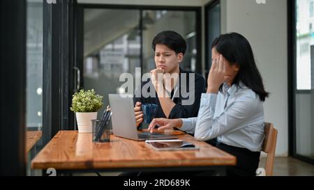 Two serious and stressed Asian office workers are working remotely at a coffee shop together, discussing work planning strategies of a project and foc Stock Photo