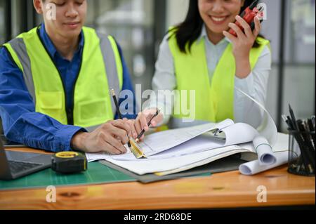 Two professional Asian construction engineers wearing safety vests are checking a blueprint, discussing work, and working in the office together. Stock Photo