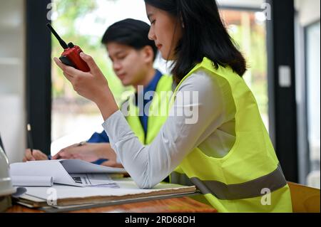 Close-up image of a professional Asian female engineer using a walkie-talkie to communicate and monitor her team while she is working in the office. c Stock Photo