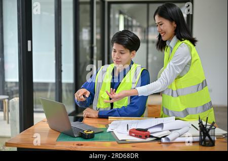 A smart and professional Asian male engineer in a safety vest is looking at a laptop screen and discussing a building blueprint on the desktop with hi Stock Photo