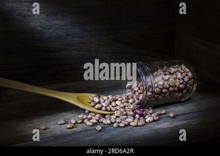 A still life of a pile of raw, dry, uncooked, beige, brown Borlotti beans spilling from a glass jar with wooden spoon in the corner of a wooden box Stock Photo
