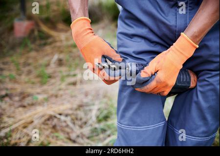 Cropped image of male worker fixing bit in electric drill. Man working on construction zone. Male builder wearing blue workwear and orange working gloves. Professional with electrical instrument. Stock Photo