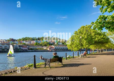 10 May 2023: Bristol, UK - A beautiful evening at Bristol Docks, young couple sitting on a bench, colourful houses,fresh green trees, blue sky, sail b Stock Photo