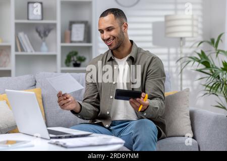 Smiling young hispanic man working remotely from home on laptop. Holds documents, reports, accounts and a calculator. Satisfied with the financial result. Stock Photo