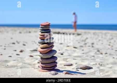 Balanced pyramid of stones on sandy beach against backdrop of bustle of woman collecting stones on seashore. Stock Photo