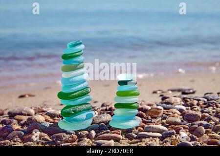 Two balanced pyramids of sea pebbles polished blue and green bottle glass against background of oncoming sea wave. Stock Photo