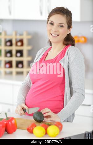 young pregnant woman holding halves of avocado in kitchen Stock Photo