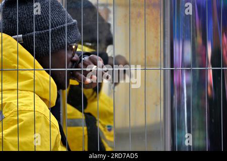 Man in yellow waterproof coat with hat behind crowed control wire fence with reflections behind Stock Photo