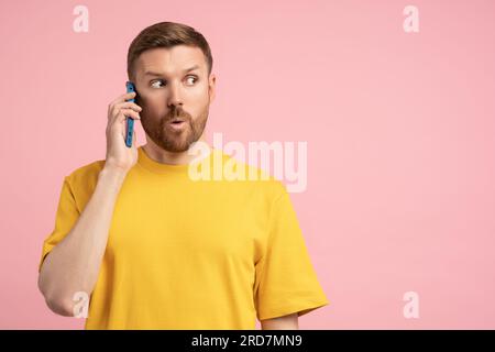 Shocked man holding cellphone surprised pointing wow eyes at copy space over pink studio background Stock Photo