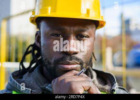 close-up portrait of young black man with beard engineer wearing yellow safety helmet, pensive with hand on chin, looking at camera standing outdoors. Stock Photo