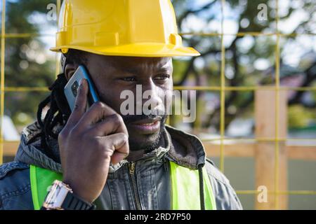 closeup portrait of young black man builder in yellow helmet, at construction site talking on the phone and looking around. Stock Photo