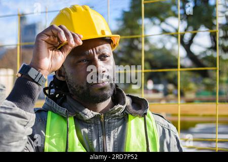 portrait of builder black man with beard wearing yellow safety helmet pensive at construction site, supervising construction process. Stock Photo