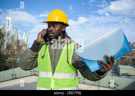 young black man civil engineer builder wearing safety helmet and reflective vest, outdoors talking on phone discussing angry with employee, holding fo Stock Photo