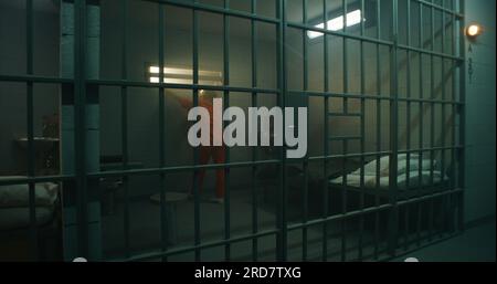 Female prisoner in orange uniform walks and stands behind metal bars, looks at barred window in jail cell. Woman serves imprisonment term for crime in prison. Depressed criminal in detention center. Stock Photo