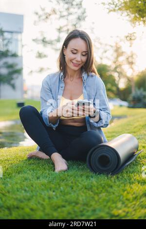 Cheerful adult woman in sportive clothes sitting on green grass field and using mobile phone. Yogi woman resting with mobile phone. Stock Photo