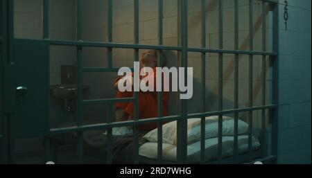 Female prisoner in orange uniform sitting on the bed in jail cell, looking at barred window. Woman serves imprisonment term for crime in prison. Depressed criminal in detention center. Stock Photo