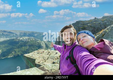 Couple taking a selfie on a rock and admiring a view on Preikestolen. Aerial shot, upper perspective on the couple. Endless view of the fjord. Stock Photo