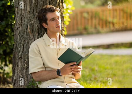Young guy sitting under tree at garden, reading book Stock Photo