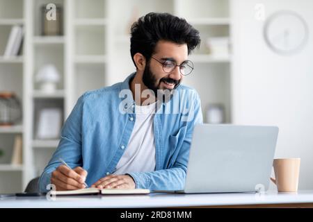 Online Lesson. Smiling Young Indian Man Using Laptop And Taking Notes Stock Photo
