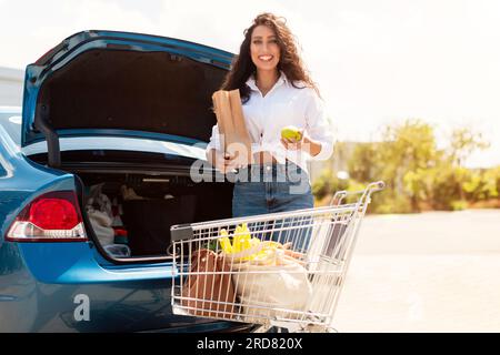 Great sales in supermarket. Happy arab woman packing grocery bags in car trunk, standing at shopping mall parking lot Stock Photo