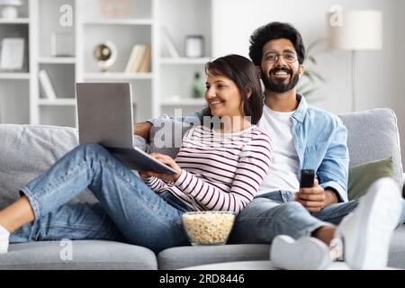 Relaxed eastern couple resting at home, watching tv, using laptop Stock Photo