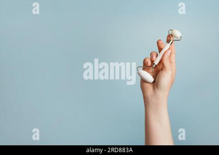 Hand holding white facial jade massage roller, gua sha on blue background. Skin care, copy space Stock Photo