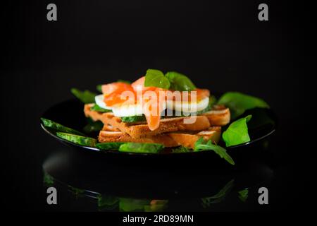 Cooked sandwich with lightly salted red fish, cucumbers and boiled eggs with herbs, isolated on a black background. Stock Photo
