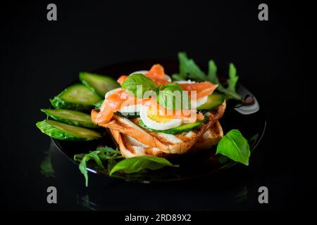 Cooked sandwich with lightly salted red fish, cucumbers and boiled eggs with herbs, isolated on a black background. Stock Photo