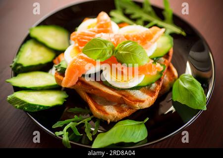 Cooked sandwich with lightly salted red fish, cucumbers and boiled eggs with herbs, in a plate. Stock Photo