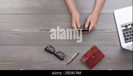 Bird's eye view of a young individual using a smartphone, accompanied by a notebook, pen, glasses, and laptop on the table Stock Photo