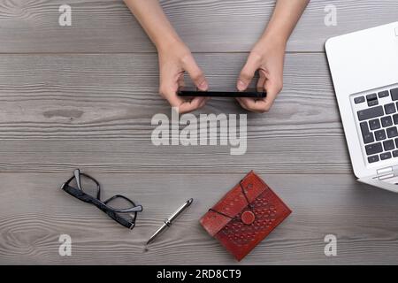 Top view of a table surface, young unidentifiable person's hands using a smartphone for work or leisure. Notebook, pen, glasses, and a laptop present Stock Photo