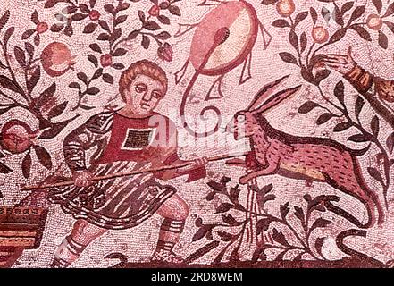 This photo of a mosaic showing a hunting scene (scene of a young man/boy hunting a hare) was taken in the summer of 1970 at Piazza Armerina in Sicily. Piazza Armerina is home to the Roman Villa del Casale and its famous mosaics, the 'finest mosaics in situ anywhere in the Roman world,' as described by UNESCO, which inserted it into its World Heritage list in 1997. Villa Romana was a lavish patrician residence built at the center of a huge latifundium (agricultural estate) at the end of the 4th century AD. It is thought to have belonged to a member of the Roman senatorial aristocracy, who trade Stock Photo