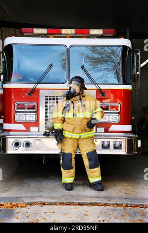 Woman firefighter stands in front of fire truck at fire station.  She is wearing bunking gear complete with gas mask and helmet. Stock Photo