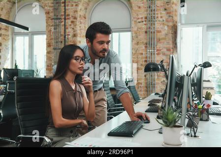 Project team. Two young colleagues in casual wear looking at computer monitor and discussing results while working in modern office Stock Photo